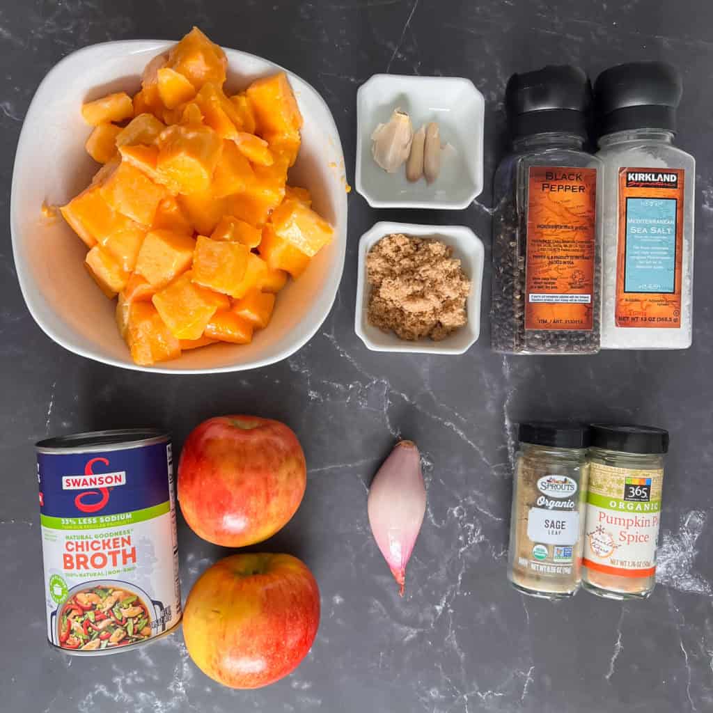 Cooked canned pumpkin chunks, garlic cloves, ginger, black pepper, sea salt, brown sugar, apples, chicken broth, sage, pumpkin pie spice, culinary spices on black marble surface for autumn or fall cooking.