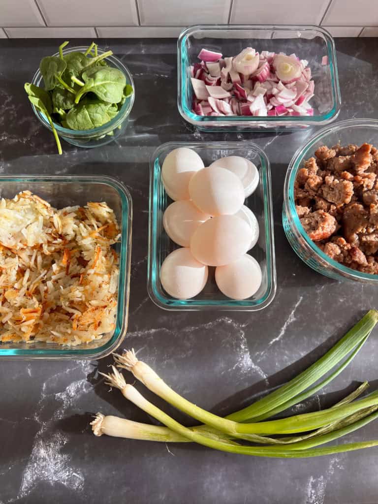 Ingredients including sausage, eggs, onion, spinach and hash browns for breakfast quiche in glass dishes 