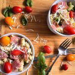 Two bowls of pasta salad on a cutting board with tomatoes, cheese and basil scattered on the table