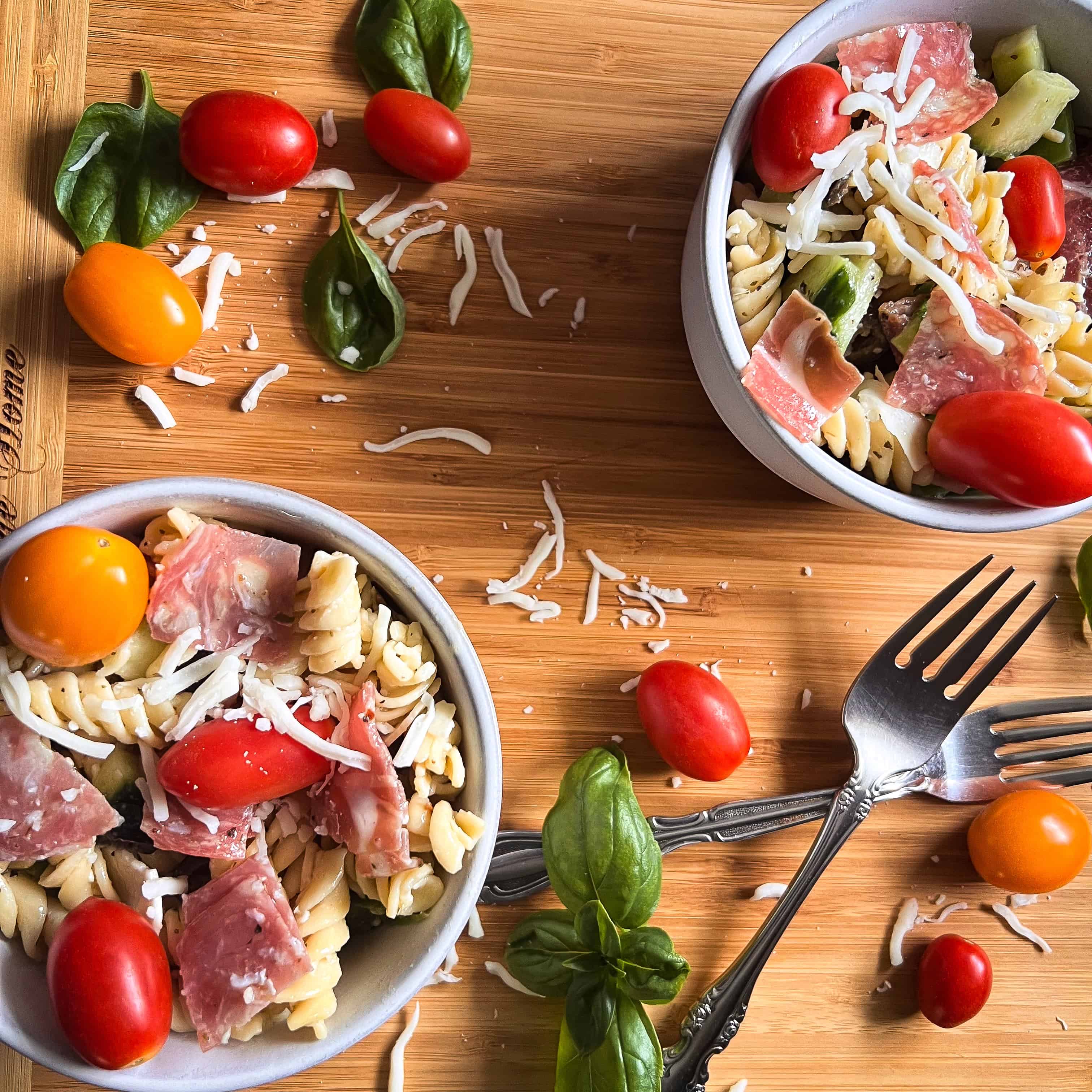 Two bowls of pasta salad on a cutting board with tomatoes, cheese and basil scattered on the table