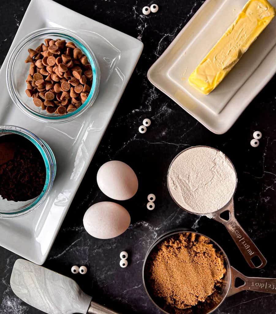 A spread of ingredients including butter, sugar, flour, cocoa powder, salted caramel chips and candy eyeballs for making Black Cocoa Mummy Brownies