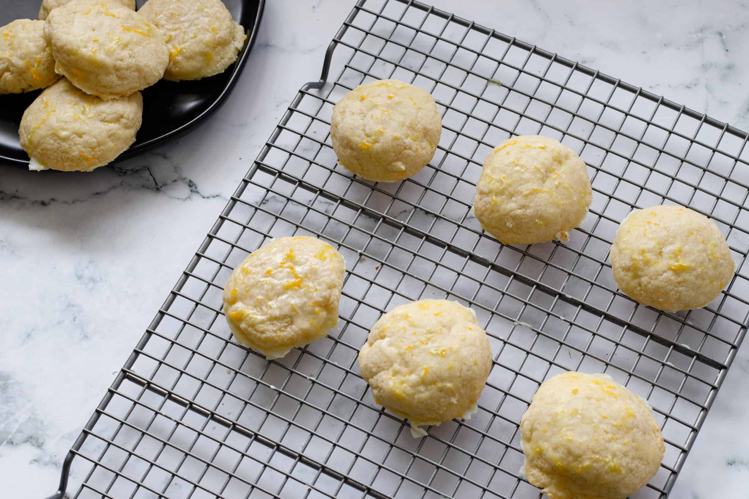 Fluffy orange glazed cookies lined up on a wire baking rack with a black plate of them beside.