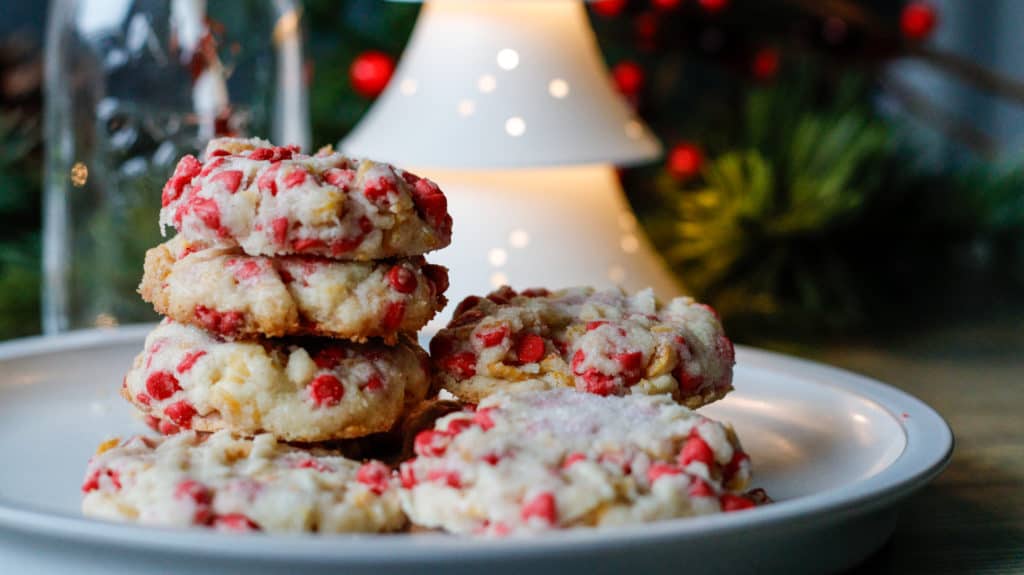 Festive shortbread base cookies with bright red cherry chips on a plate with holiday decor in the background