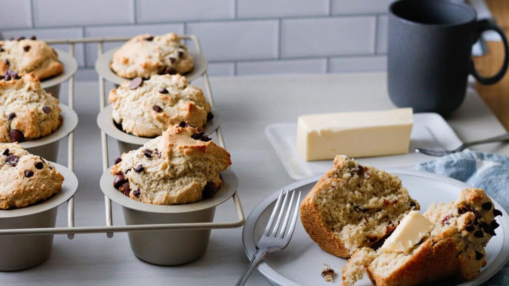 Chocolate chip muffin a plate, with muffins in tin behind it and a stick of butter and coffee mug in the background.