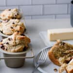 Chocolate chip muffin a plate, with muffins in tin behind it and a stick of butter and coffee mug in the background.