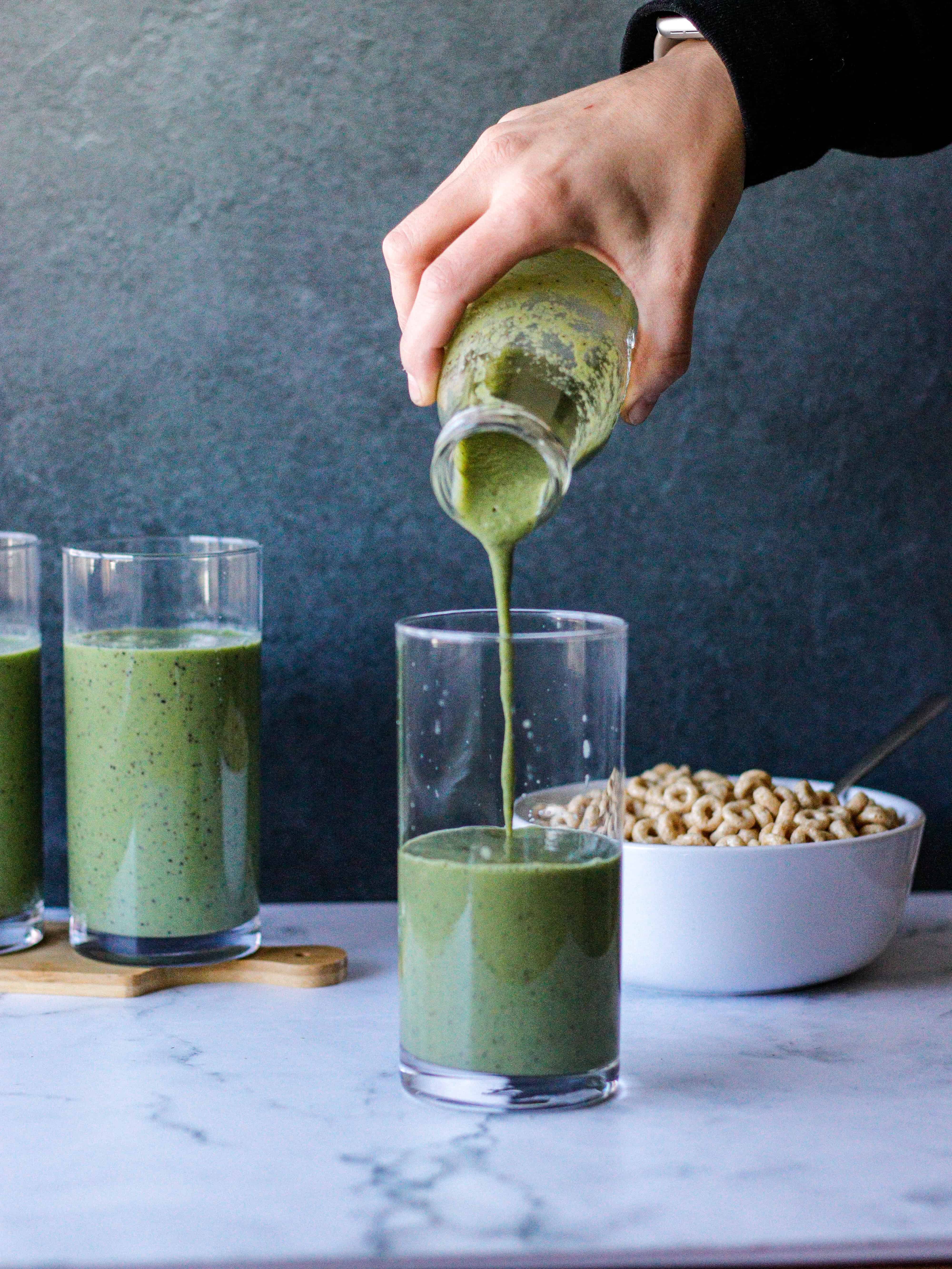 Pouring a green smoothie into a glass with bowl of cereal in the background