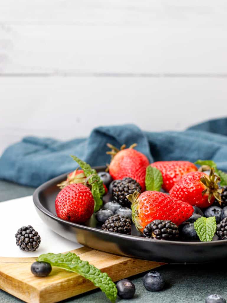 Fresh mixed berries including strawberries, blackberries, and blueberries served in a black bowl on a wooden board.