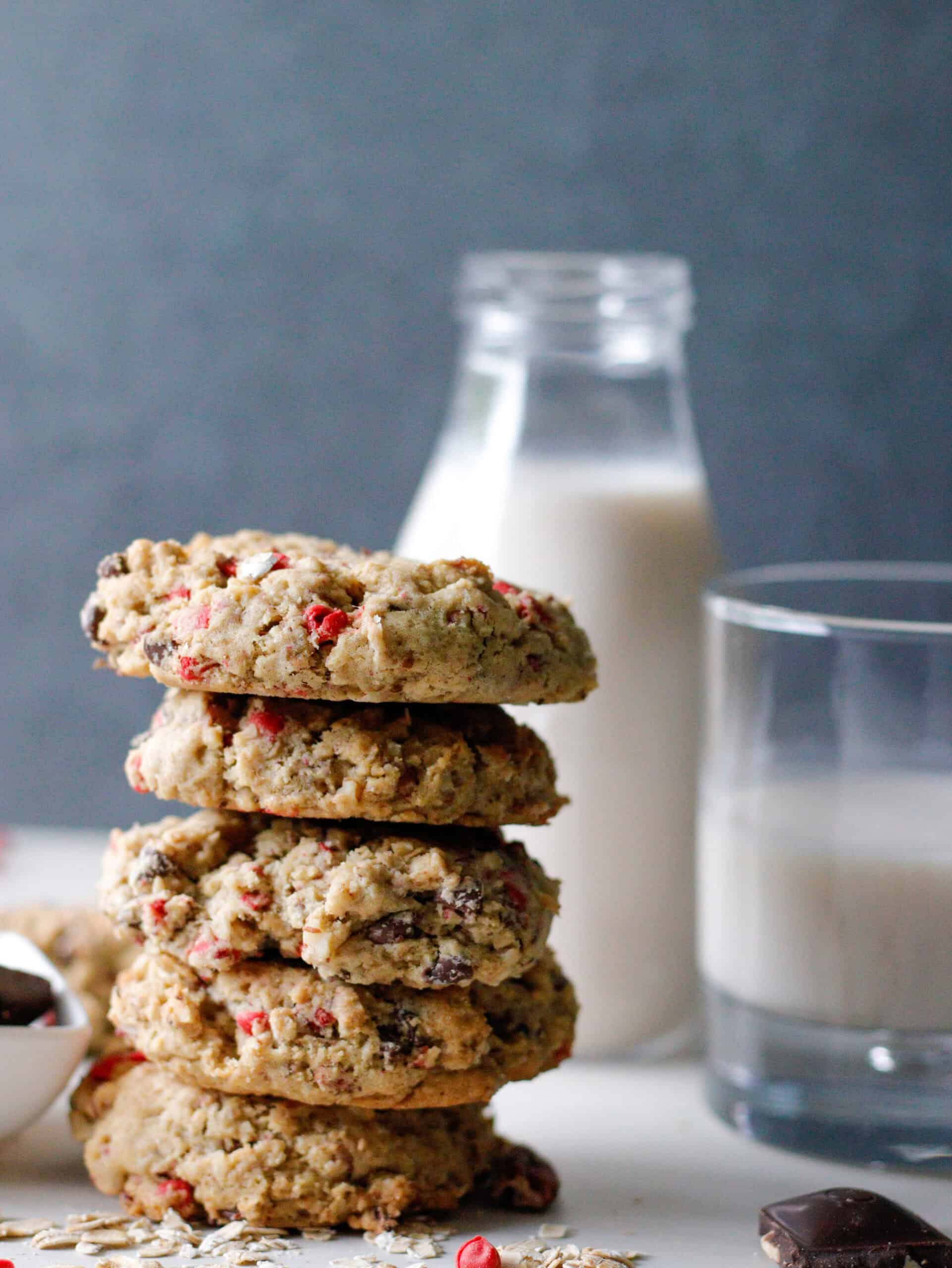 Crispy oatmeal cookies with red peppermint bits, glass of milk, and chocolate chunks on white surface.