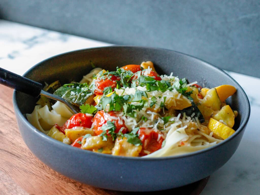 Fresh vegetable pasta dish with cherry tomatoes, zucchini, and fresh herbs in a black bowl on a wooden surface.