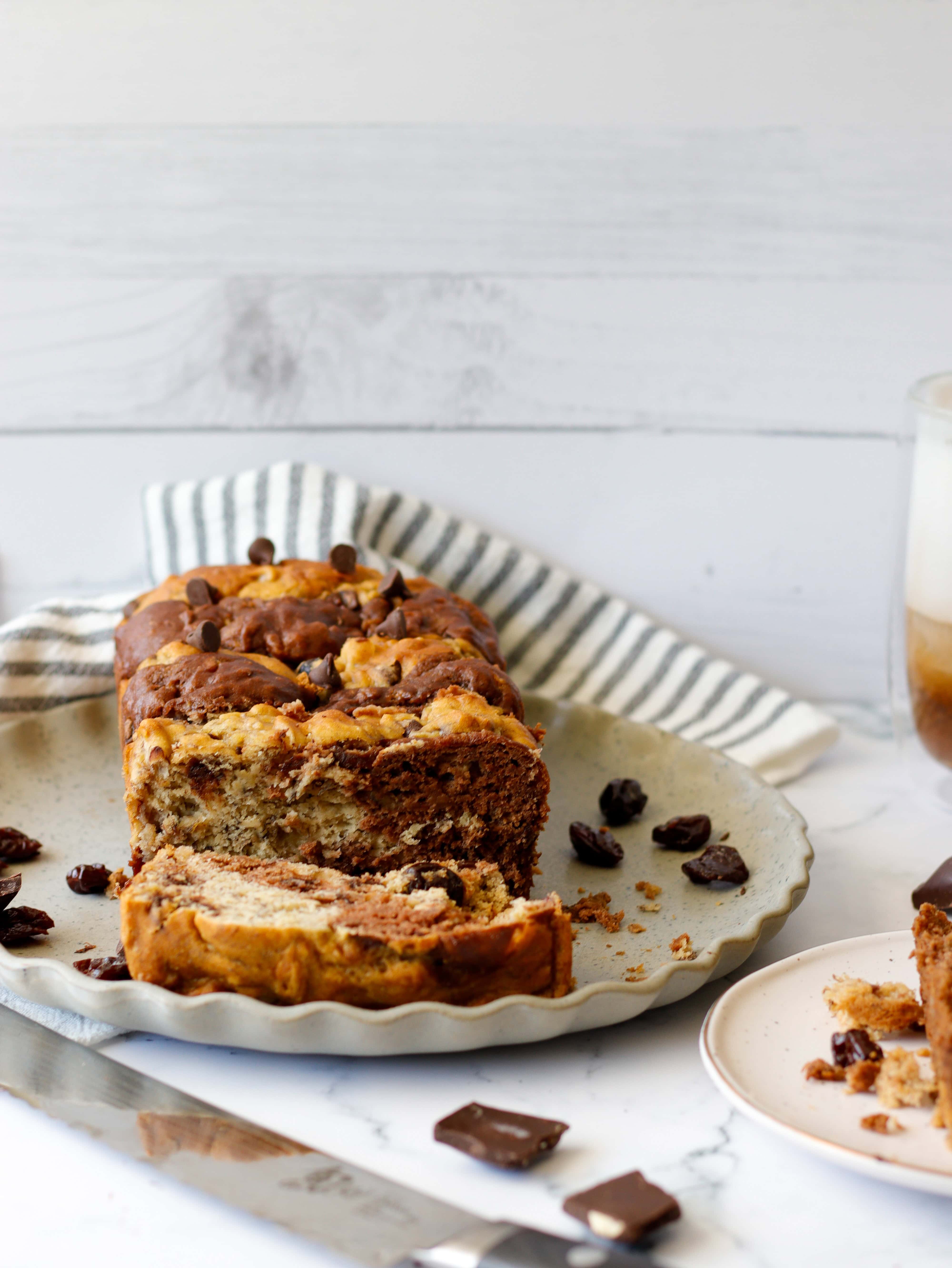 Baked dessert bread on a plate with a bread knife, chocolate chips and coffee in the background.