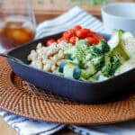 Salad with beans, broccoli, zucchini, tomatoes in a dark square bowl with a glass of iced tea in the background.