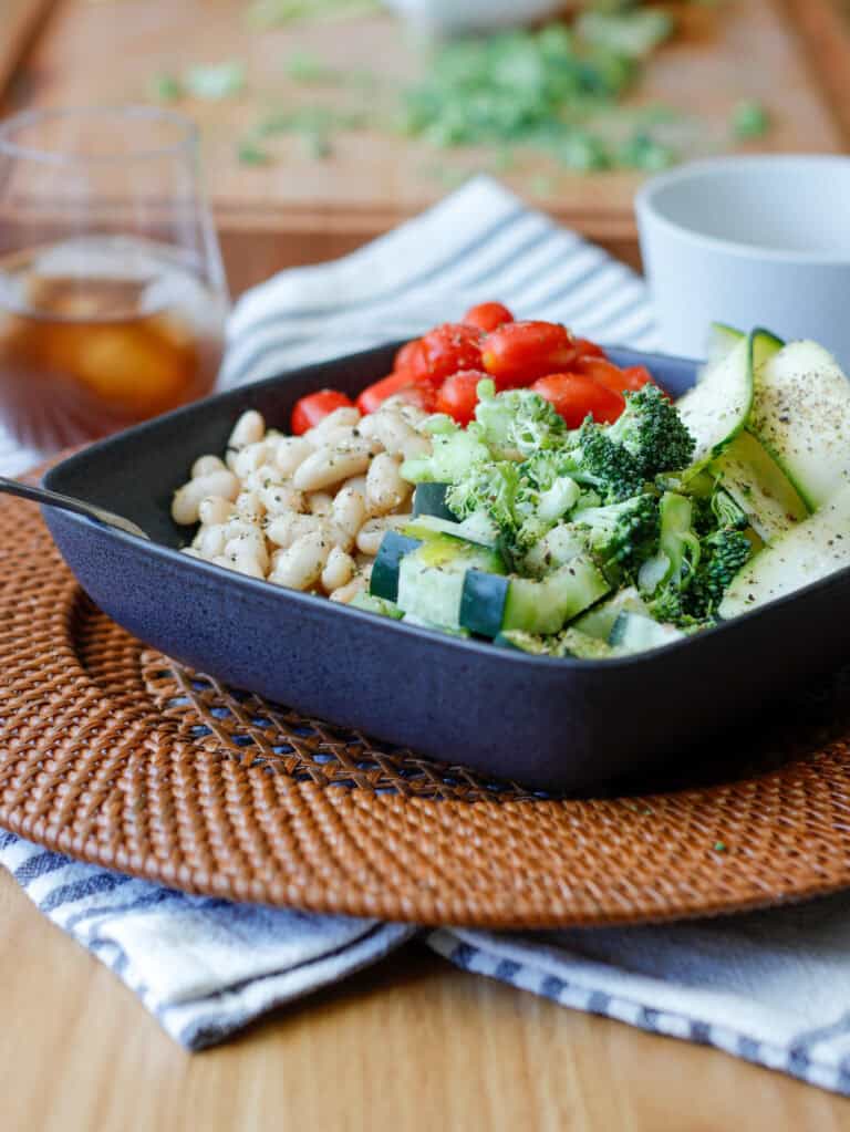 Salad with beans, broccoli, zucchini, tomatoes in a dark square bowl with a glass of iced tea in the background.