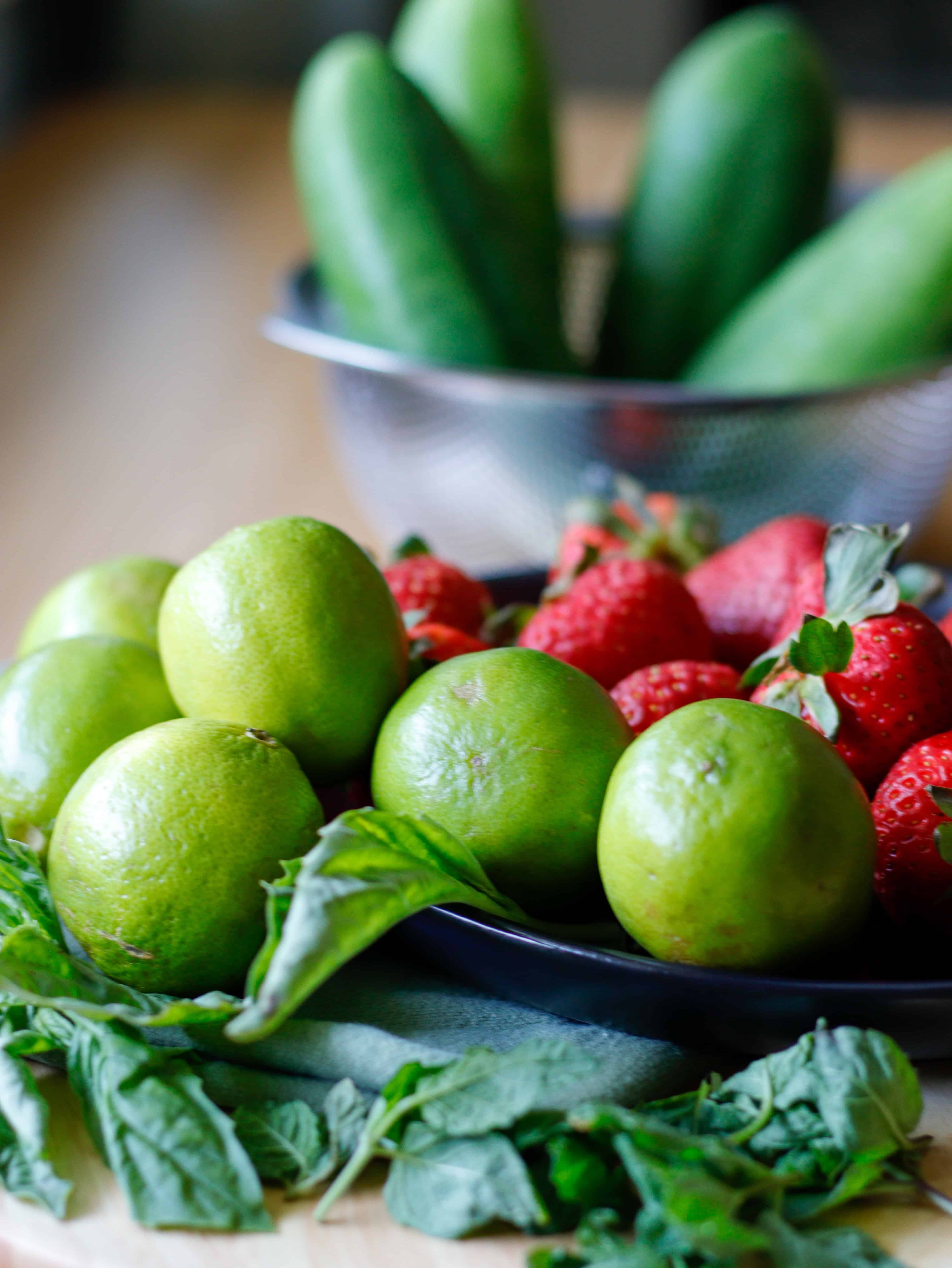 Limes on a green napkin with mint, strawberries and cucumbers in the background