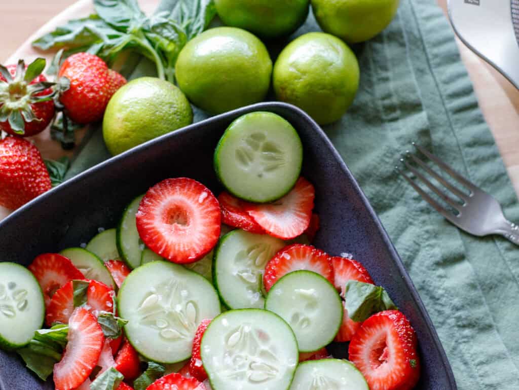 Close up of sliced cucumbers and strawberries in a bowl on top of a green dinner napkin with limes next to the bowl