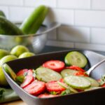 Bowl with sliced strawberries and cucumbers with cucumbers in a bowl in the background.