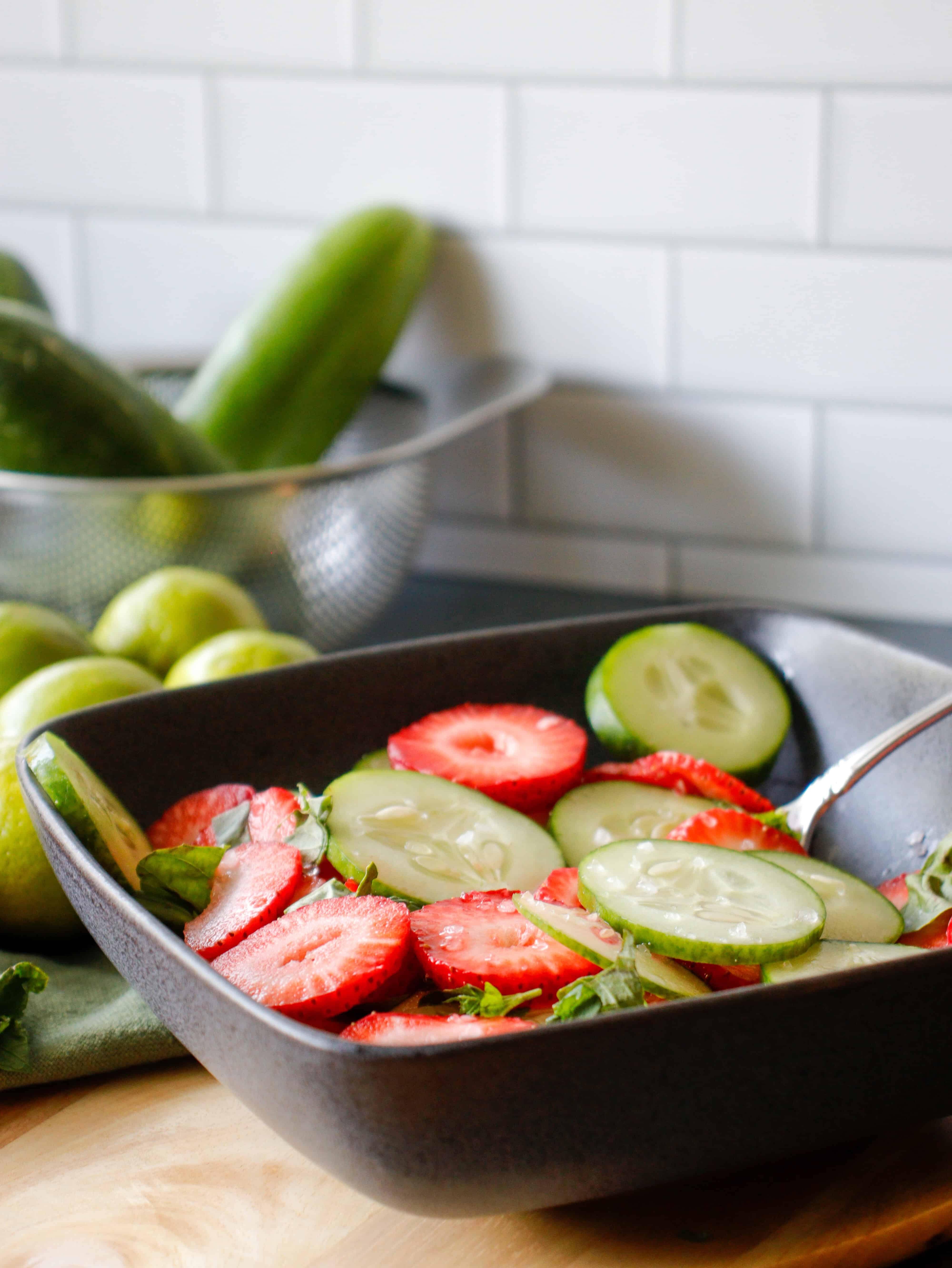 Bowl with sliced strawberries and cucumbers with cucumbers in a bowl in the background.