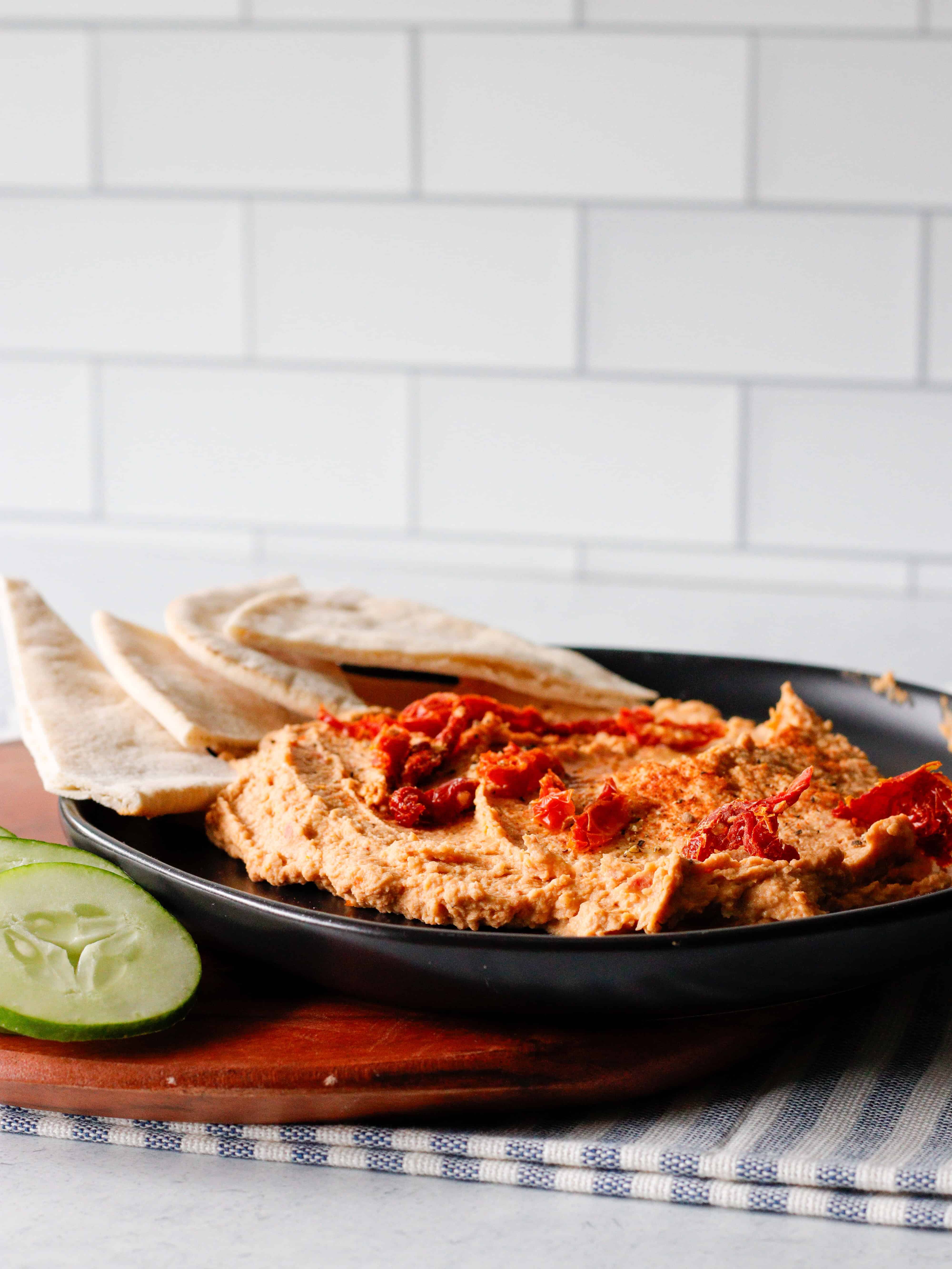 Black plate with sun-dried tomato hummus and pita bread in the background.
