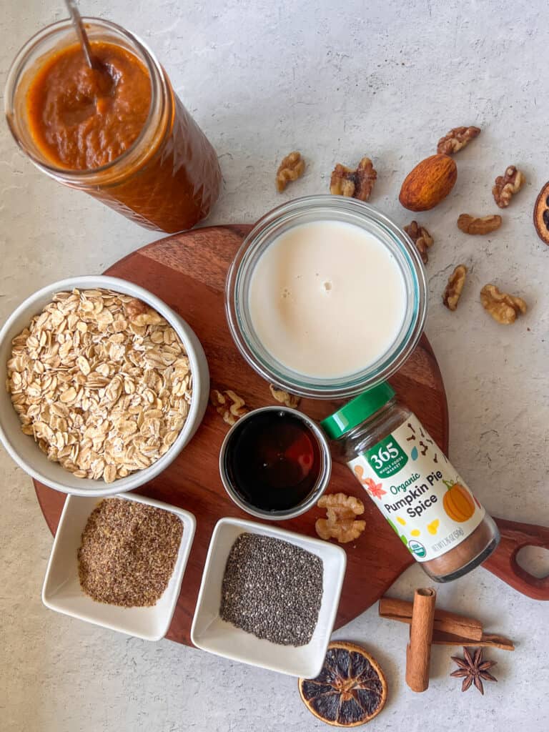 A layout of ingredients for pumpkin pie overnight oats on a table. Chia seeds, flax seed, oats, pumpkin butter, almond milk and cinnamon.