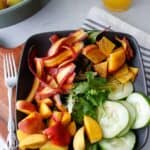 Bowl filled with cucumbers, salad greens, purple carrots and peaches on a stripped napkin with peaches in the background.