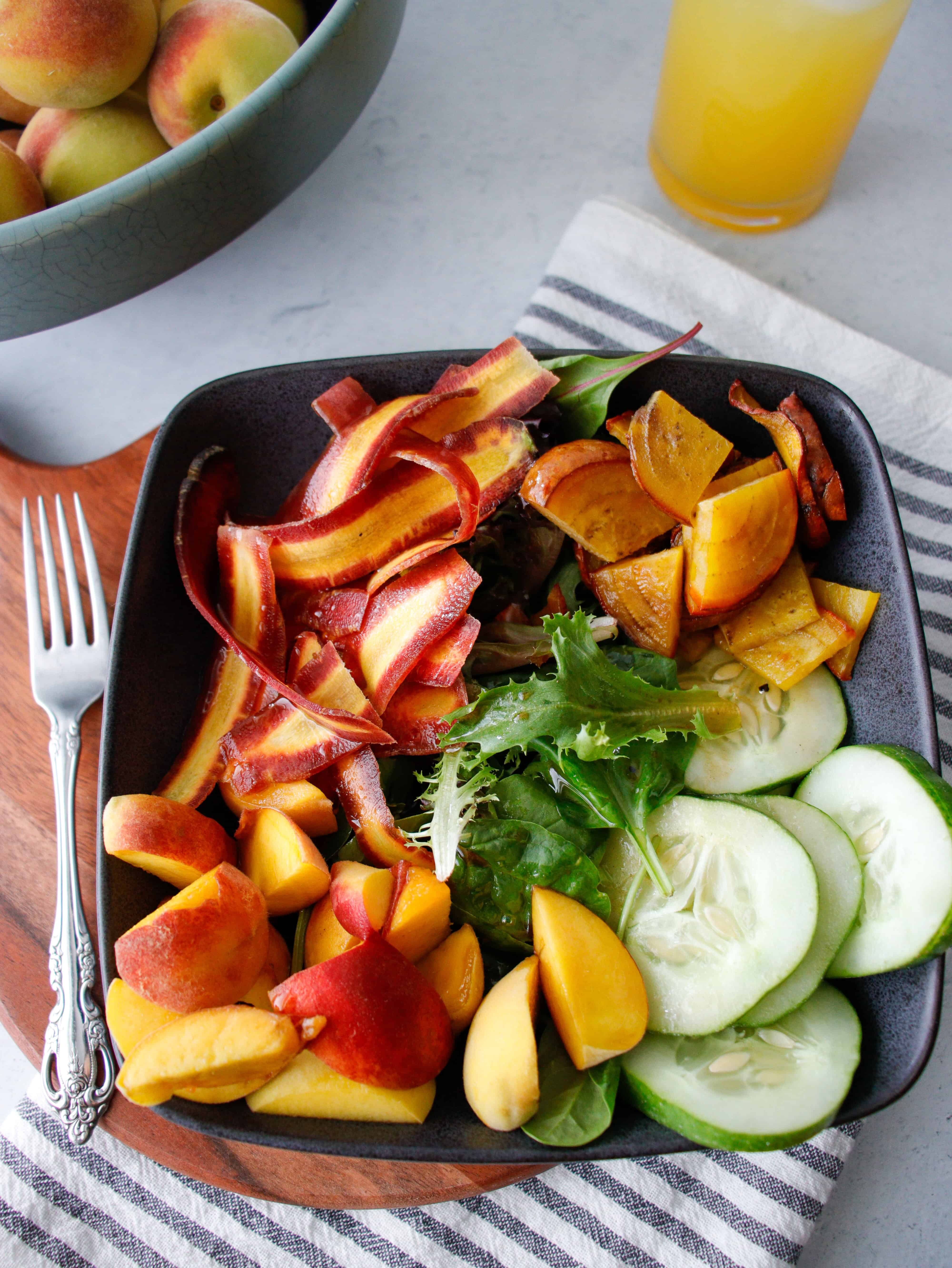 Bowl filled with cucumbers, salad greens, purple carrots and peaches on a stripped napkin with peaches in the background.