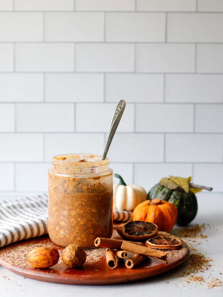 Pumpkin Pie flavored overnight oats in a jar with a spoon on a wood cutting board with pumpkins in the background and cinnamon sticks