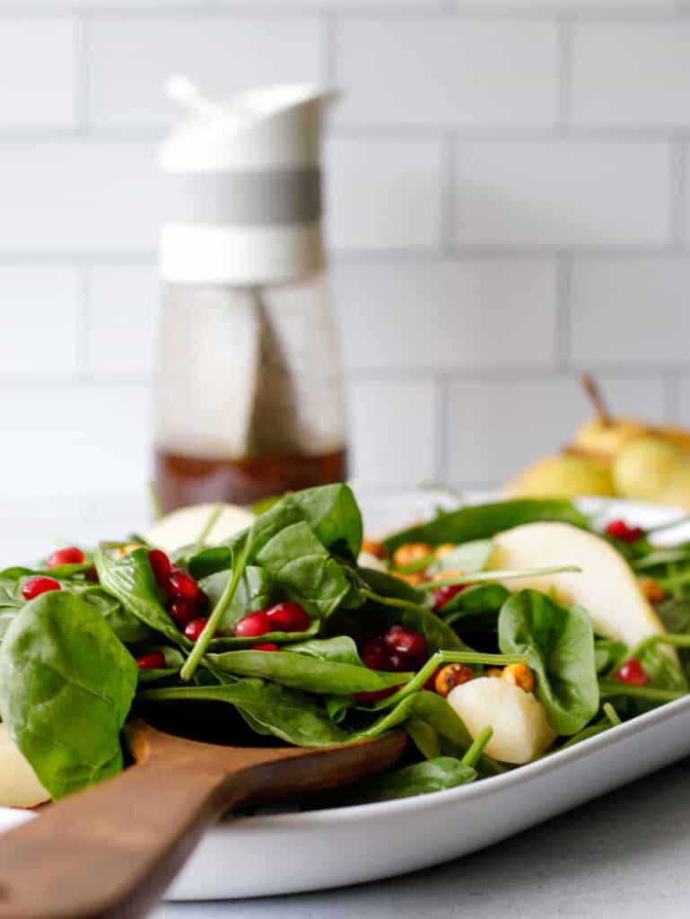 Spinach salad on a long white plate with pears and pomegranates on top and a dressing container in the background.