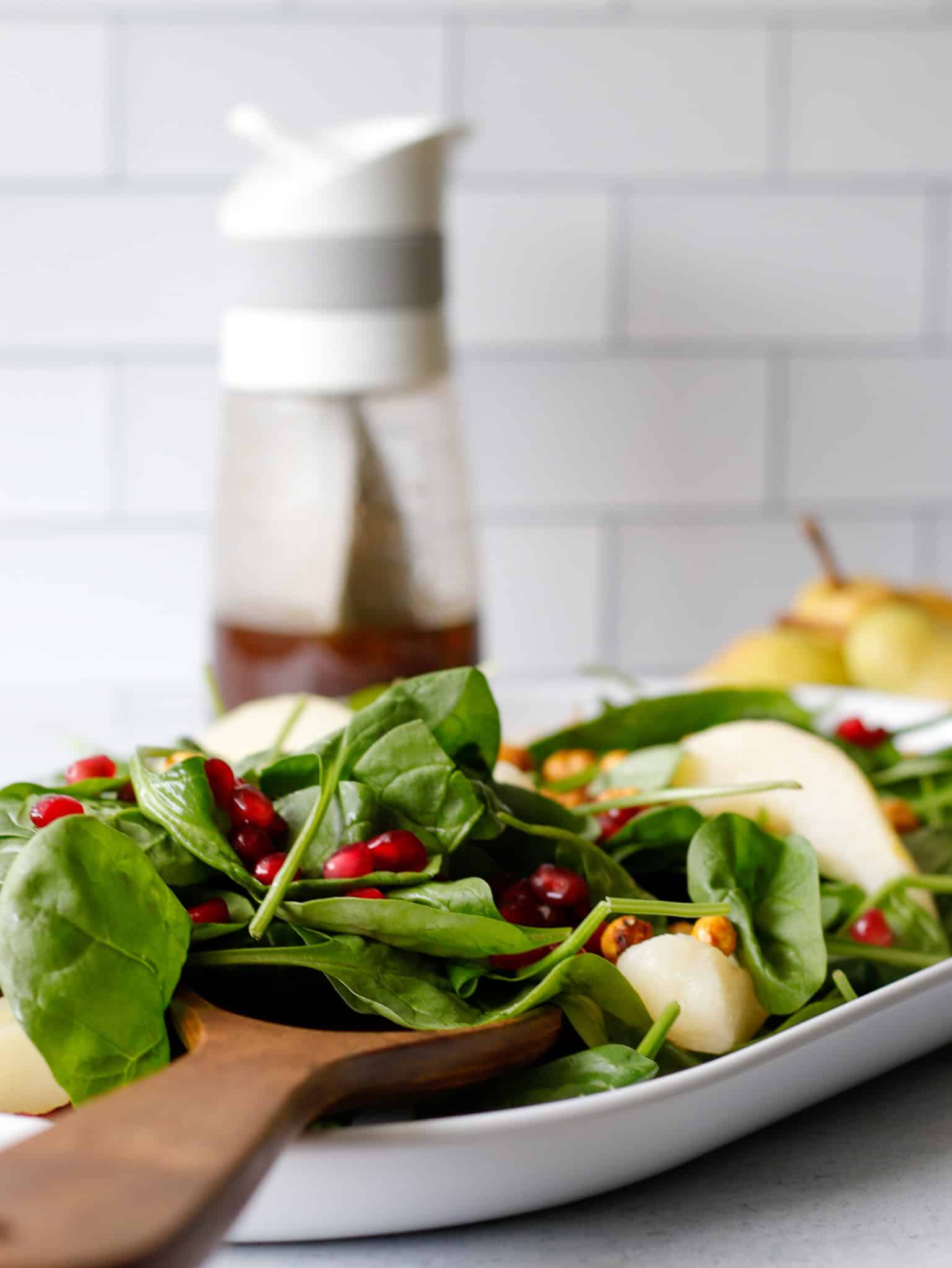 Spinach salad on a long white plate with pears and pomegranates on top and a dressing container in the background.