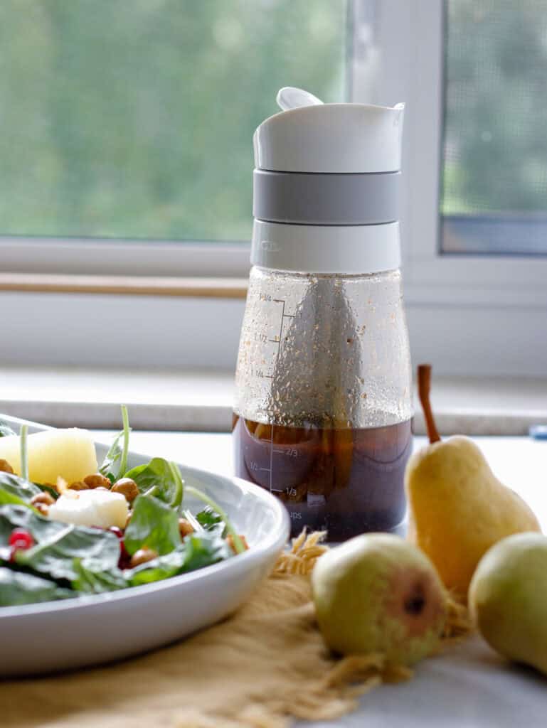A plated salad, pears and a container of dressing sitting in front of a window. 