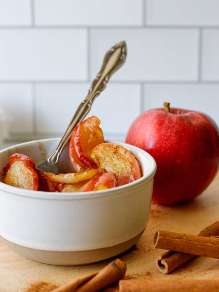 Cinnamon apples in a small white bowl with a fork and a full gala apple in the background.