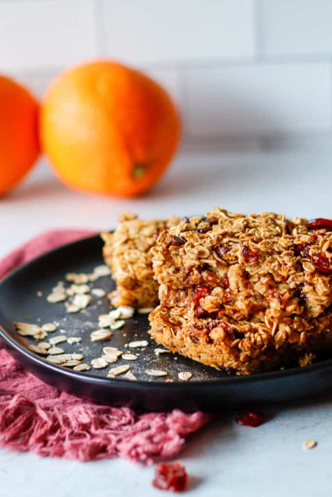 Stacked simple oatmeal bars on a black plate with oranges in the background.