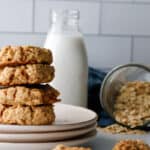 Oatmeal cookies stacked on a white plate with milk and a mason jar of oats spilled over in the background