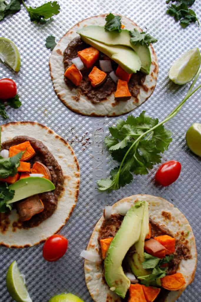 Three tacos, with crispy tortillas on a baking dish topped with black bean, sweet potato, avocado and cilantro
