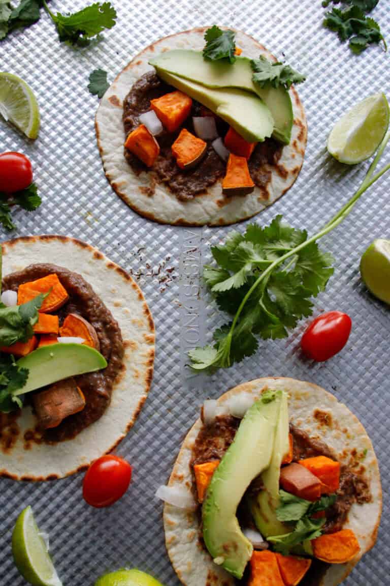 Three tacos, with crispy tortillas on a baking dish topped with black bean, sweet potato, avocado and cilantro