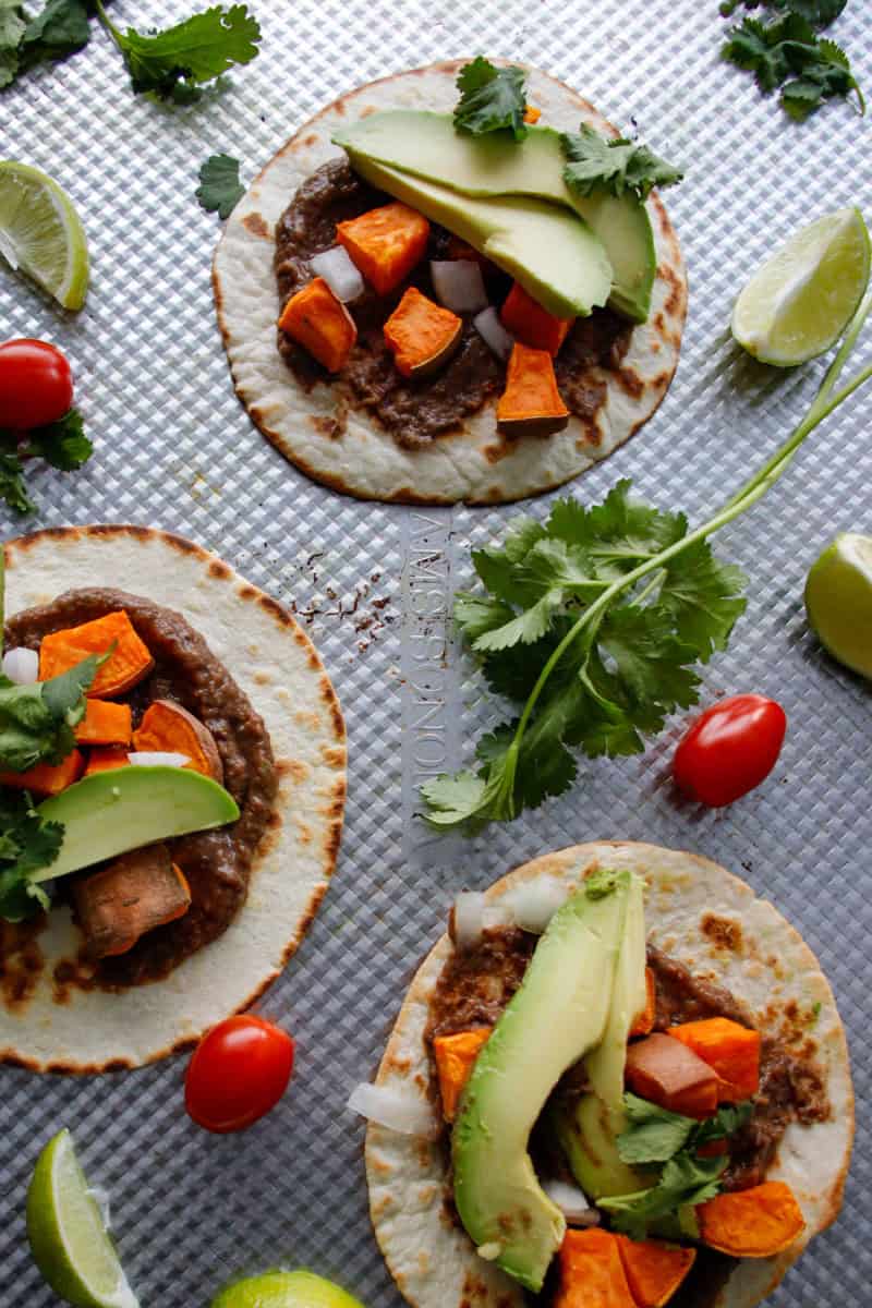Three tacos, with crispy tortillas on a baking dish topped with black bean, sweet potato, avocado and cilantro