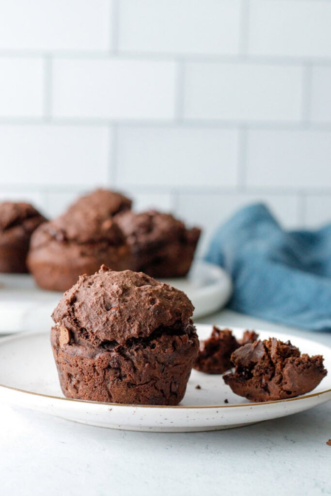 Gluten free chocolate espresso muffin on a white plate with piece broken off and a full tray in the background