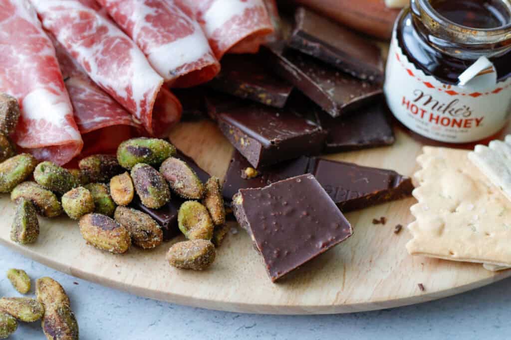 Cheese board focused on pieces of chocolate, cinnamon pistachios, cured meat and mike's hot honey