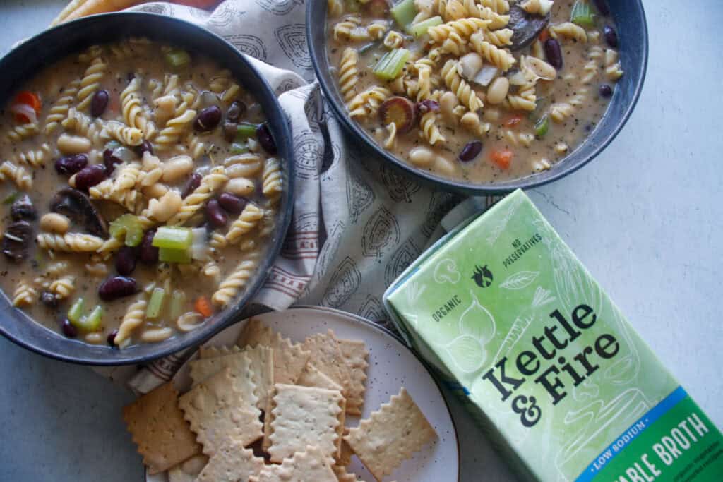 two bowls of vegan chickpea noodle soup with a side of crackers and a kettle and fire broth carton