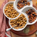 Overhead of crispy chickpeas in a four section bowl on a brown cutting board, with a napkin and seasoning bottles scattered.