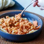 Picture of a hand with a fork eating creamy cauliflower pasta sauce over rotini noodles in a gray bowl with sliced bread in the background.