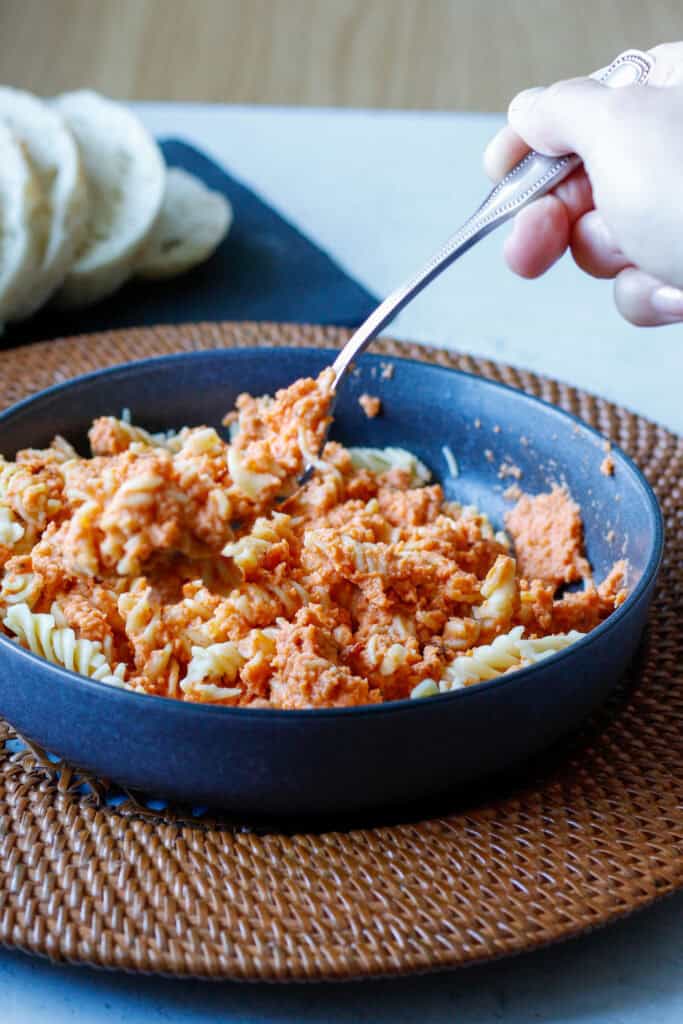 Picture of a hand with a fork eating creamy cauliflower pasta sauce over rotini noodles in a gray bowl with sliced bread in the background.