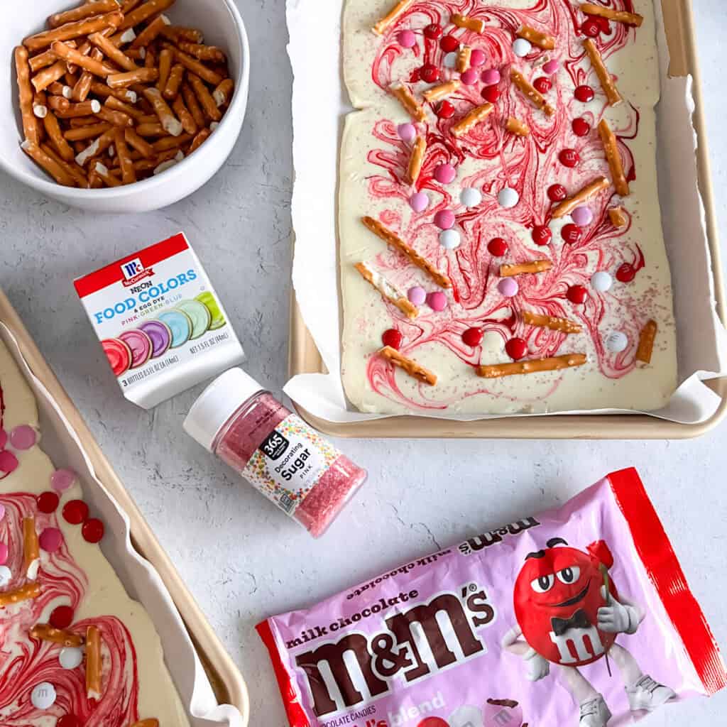 Overhead look of white chocolate bark and the ingredients including pretzels, sugar, M&Ms and food coloring