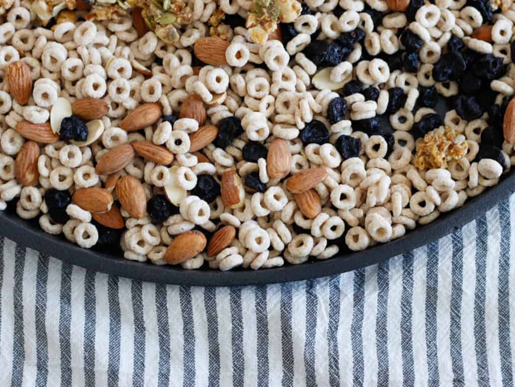 Almonds, blueberries, and toasted cereal mix in a black tray on a striped cloth background.