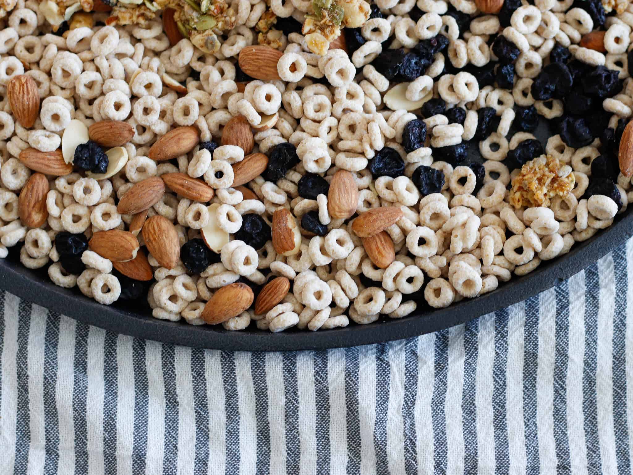 Almonds, blueberries, and toasted cereal mix in a black tray on a striped cloth background.